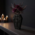 Schwarze Gothic-Sargvase mit Kreuz-Totenkopf-Relief und dunkelroten Rosen auf dunklem Sideboard, Frontalansicht mit Kerzenlicht im Hintergrund