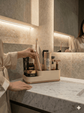 Beige-goldener Badezimmer-Organizer auf Marmor-Waschtisch, befüllt mit elektrischer Zahnbürste, Cremetiegel und mehreren Pflegeprodukt-Flaschen, Hand greift nach der Zahnbürste, im Hintergrund Person im Seidenmantel und beleuchteter Spiegel.