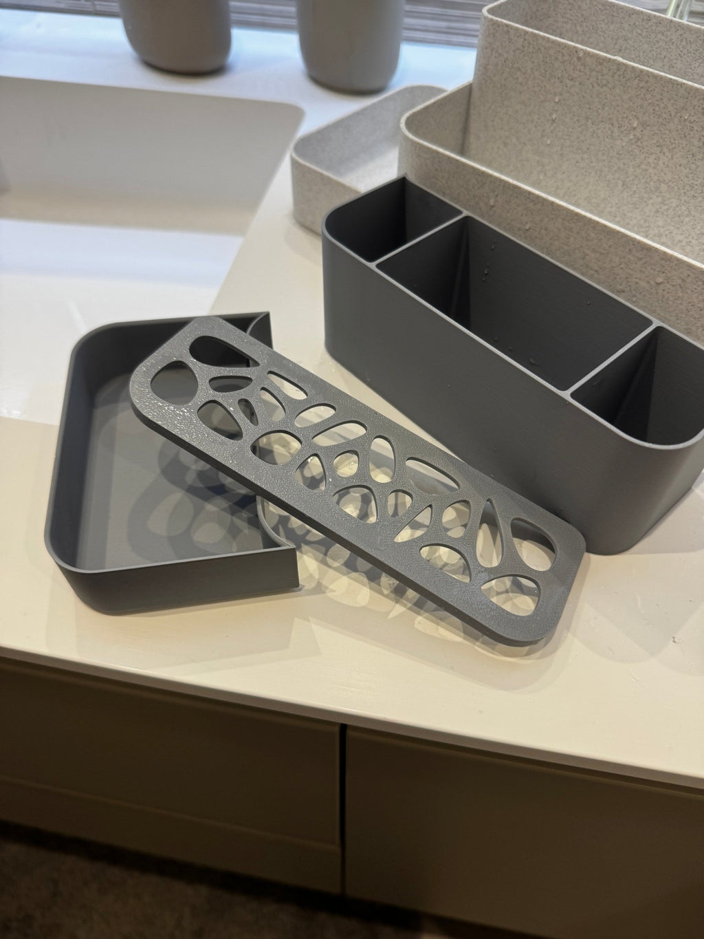 Zerlegte Innenteile des Badezimmer-Organizers, echtes Foto: dunkelgraue Ablaufschale, perforierte Voronoi-Abtropfgitter-Einlage und Fächer-Tray einzeln nebeneinander, im Hintergrund die hellgrauen Außenschalen unscharf, auf heller Ablage.