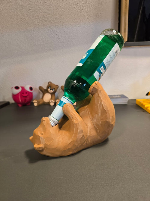 Bärenförmiger Weinflaschenhalter in beiger Wunschfarbe, Frontalansicht mit grüner Flasche im Maul, Deko-Figuren im Hintergrund – Münch3D
