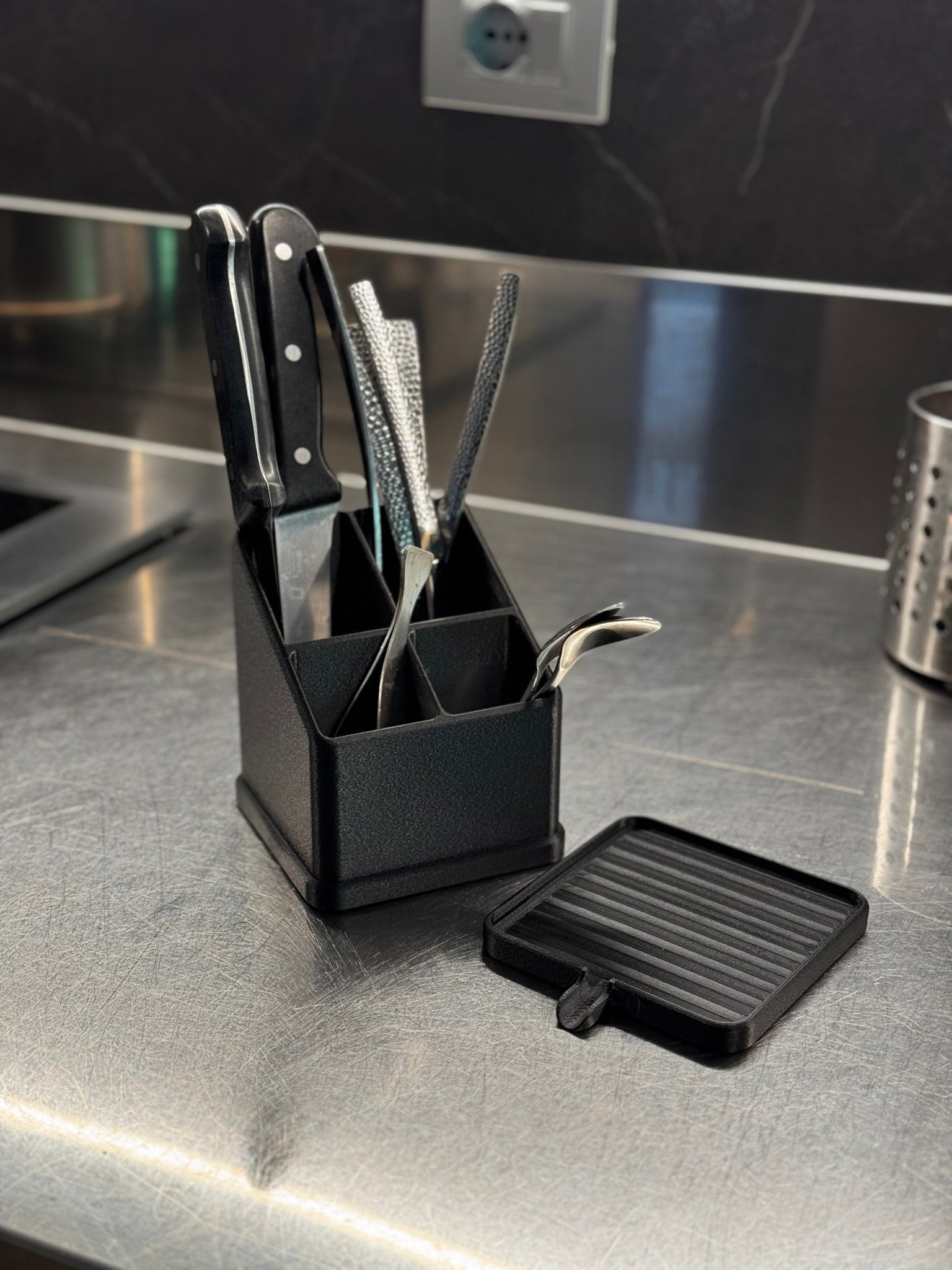 Echtfoto: Schwarzer Besteckhalter mit Messern, Reibeisen und Löffel befüllt, daneben eine separate flache Ablaufschale mit Rillenmuster, auf Edelstahl-Arbeitsfläche vor dunkler Marmorwand.