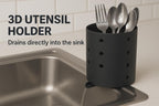 Marketing-Grafik Nahaufnahme – schwarzer perforierter Besteckhalter mit Gabel und Löffeln auf Spülenrand, Wasserstrahl läuft aus dem Ausguss in die Spüle (englischer Werbetext im Bild)