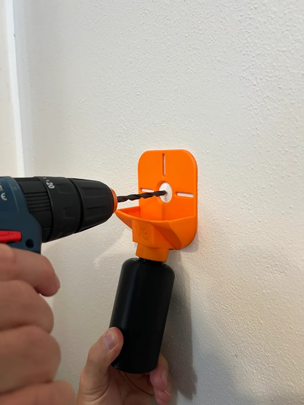 Person bohrt mit Akkuschrauber durch einen orangen Bohrstaub-Fangtrichter in eine weiße Wand, der Bohrstaub fällt in die schwarze Sammelflasche darunter