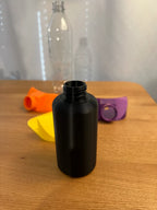 Schwarze Sammelflasche mit sichtbarem Schraubgewinde am Flaschenhals, im Hintergrund Trichteraufsätze in Orange, Gelb und Lila sowie eine PET-Flasche