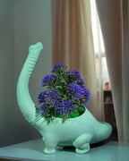 Mintgrüner Brachiosaurus-Blumentopf mit violettem Blütenstrauß, Nahaufnahme vor Vorhang und heller Wand