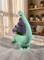 Mintgrüner Brachiosaurus-Blumentopf mit violettem Blütenstrauß, Rückansicht mit sichtbarer geriffelter Halsstruktur, auf Wolldecke mit Regal im Hintergrund