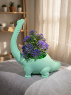 Mintgrüner Brachiosaurus-Blumentopf mit violettem Blütenstrauß, seitliche Ansicht auf heller Tischplatte, unscharfer bunter Wohnraum mit Spielzeug-Regal und Vorhang im Hintergrund