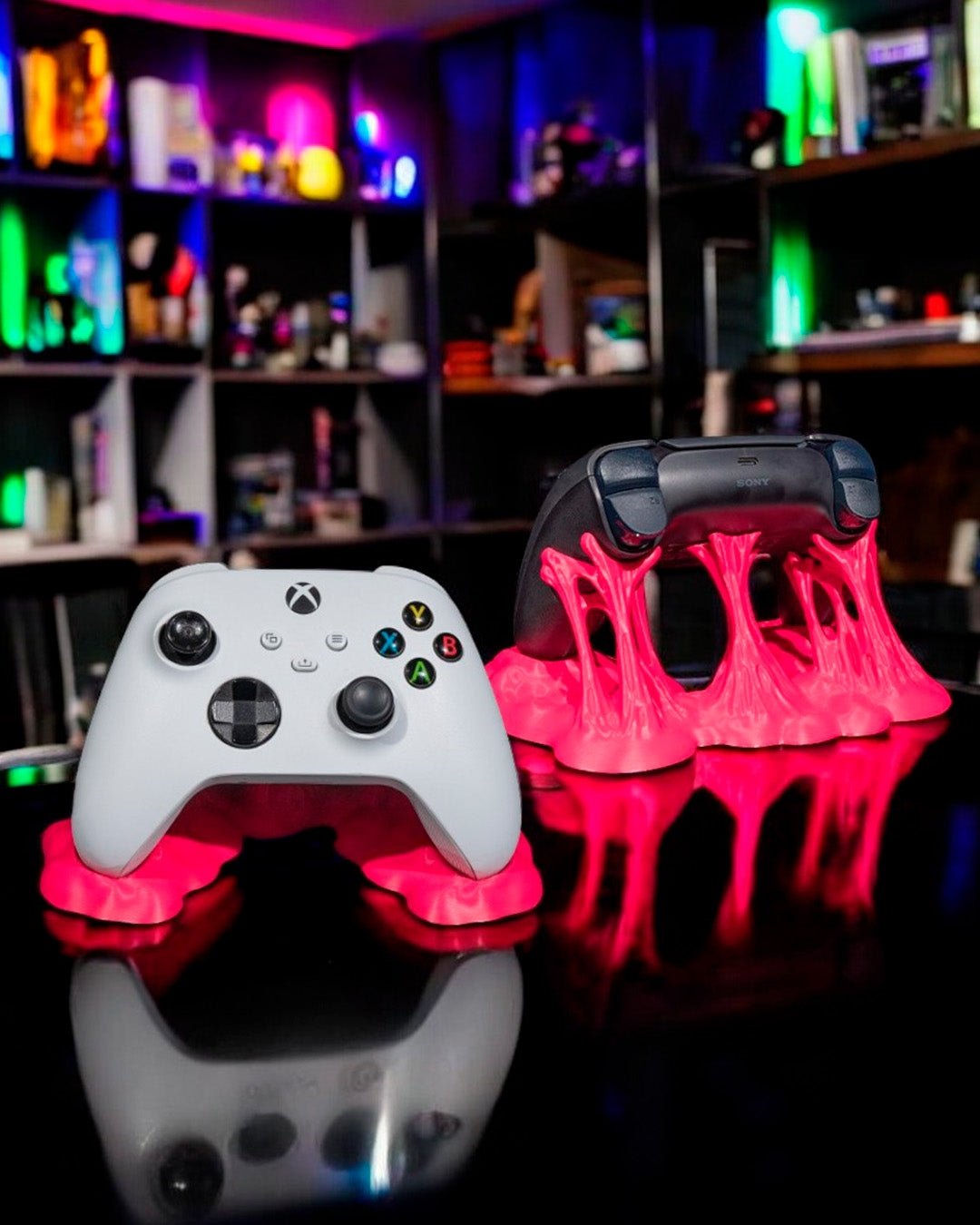 Zwei pinke Controller-Ständer im Sticky-Splash-Design auf einem Schreibtisch, im Hintergrund unscharfe Regale mit bunter Neon-Beleuchtung – links weißer, rechts schwarzer Gaming-Controller aufgestellt