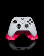 Weißer Gaming-Controller in pinkfarbenem Sticky-Splash-Controller-Ständer, Frontalansicht vor schwarzem Hintergrund