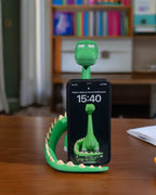 Grüner Dino-Handyhalter mit eingestecktem Smartphone und sichtbarem Sperrbildschirm, auf Holzschreibtisch mit Bücherregal im Hintergrund