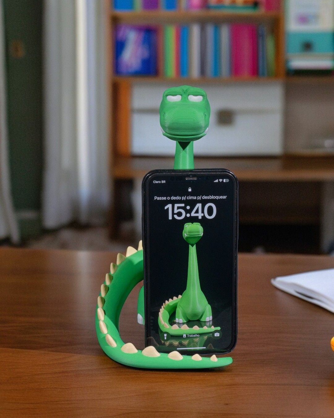 Grüner Dino-Handyhalter mit eingestecktem Smartphone und sichtbarem Sperrbildschirm, auf Holzschreibtisch mit Bücherregal im Hintergrund