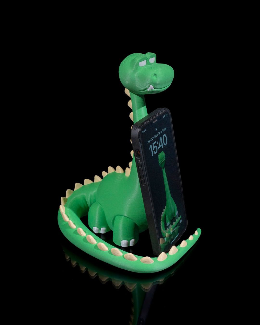 Grüner Dino-Handyhalter mit eingestecktem Smartphone, Frontalansicht aus leicht anderem Blickwinkel vor schwarzem Hintergrund