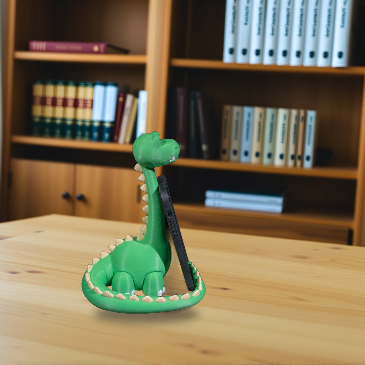 Grüner Dino-Handyhalter mit eingestecktem Smartphone, seitliche Ansicht auf hellem Holzschreibtisch mit unscharfem Bücherregal im Hintergrund