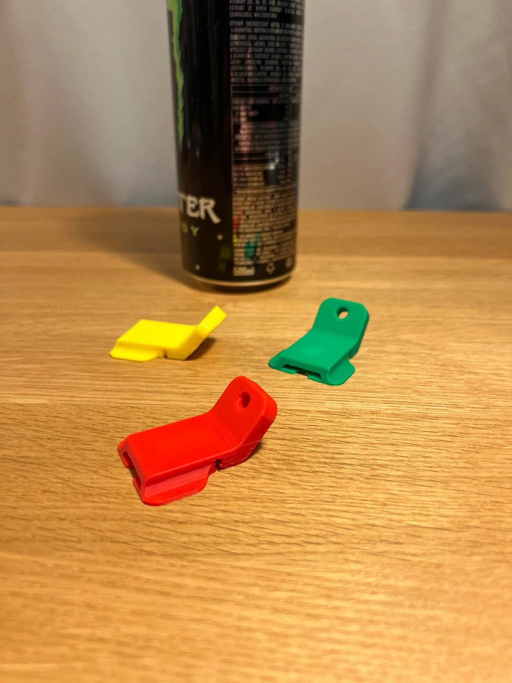 Drei Dosenöffner-Clips in Gelb, Rot und Grün auf Holztisch, Getränkedose im Hintergrund