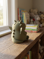 Grüne Drachen-Spardose von hinten mit Flügeln und Rückenstacheln am Fenster, Kinderbücher und Kuscheltiere im Hintergrund