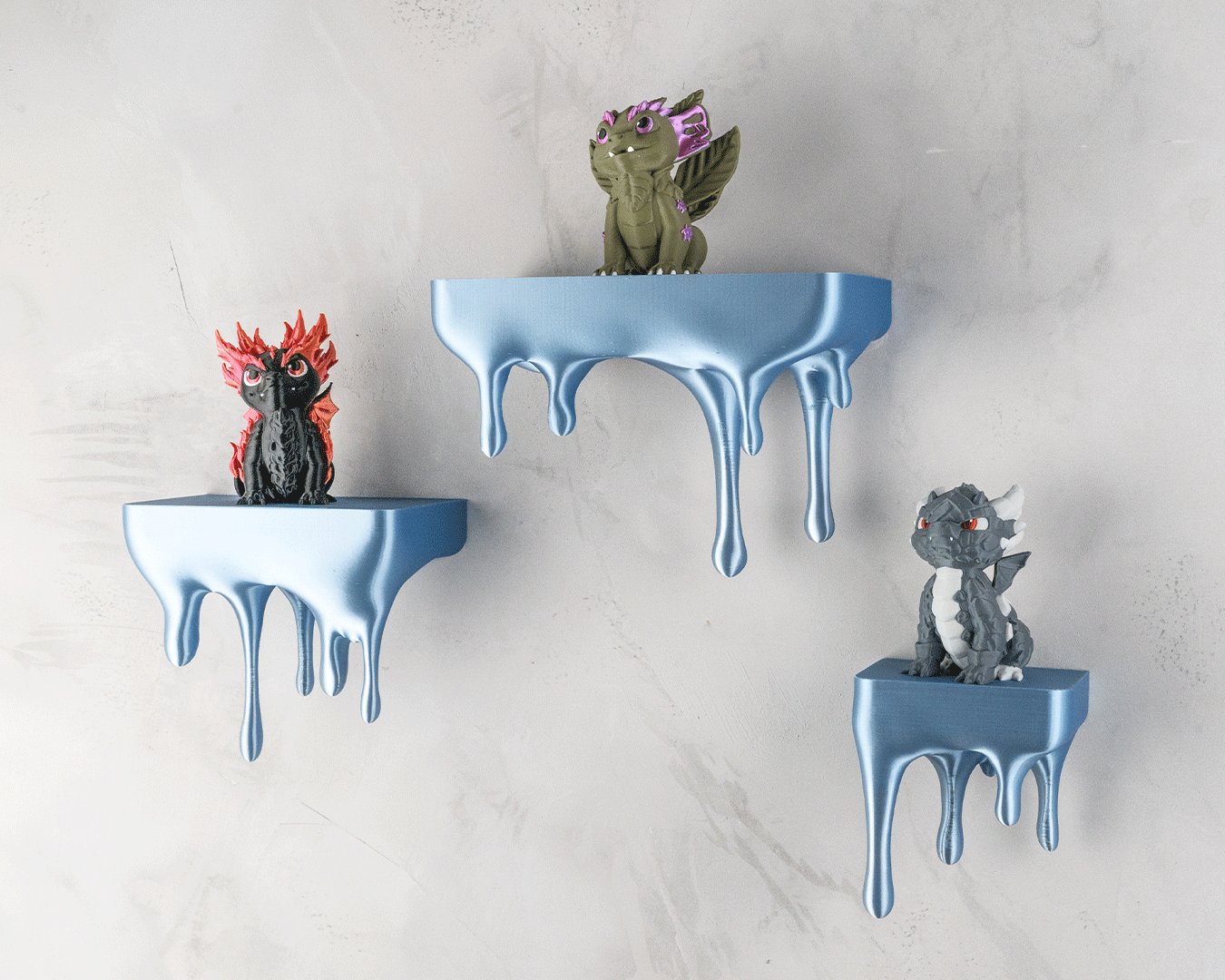 Drei blaue Tropfen-Wandregale mit Drachenfiguren (rot, oliv, grau) an heller Steinwand, Nahaufnahme