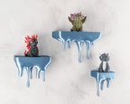Drei blaue Tropfen-Wandregale mit Drachenfiguren (rot, oliv, grau) an heller Steinwand, etwas näher herangezoomt