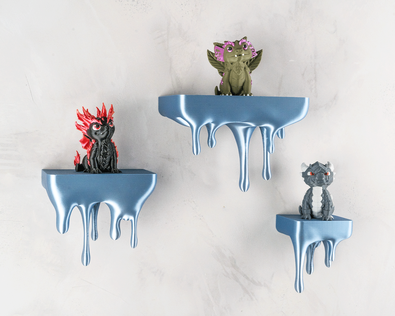 Drei blaue Tropfen-Wandregale mit Drachenfiguren (rot, oliv, grau) an heller Steinwand, etwas näher herangezoomt