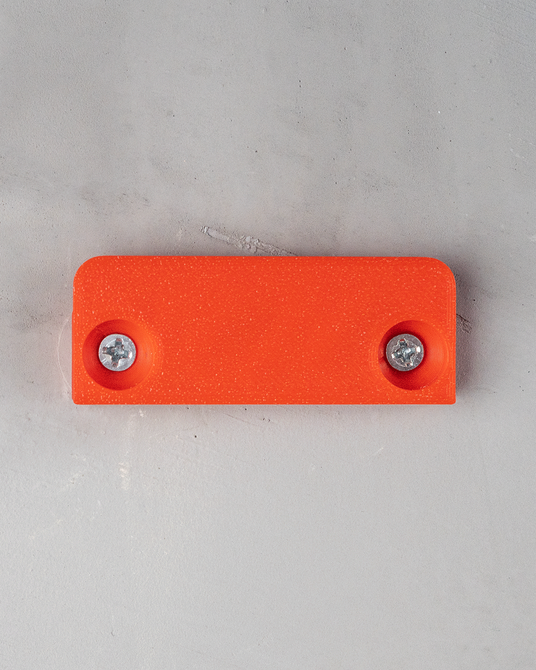 Rechteckige Montageplatte eines Tropfen-Wandregals in Orange mit zwei eingeschraubten Schrauben, Draufsicht an Wand