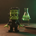 Frankenstein-Schlüsselanhänger als grüne Chibi-Figur, Frontalansicht auf einem Labortisch mit leuchtend grünem Glaskolben.