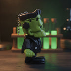 Frankenstein-Schlüsselanhänger seitlich fotografiert, im Hintergrund leuchtend grüne Glasfläschchen auf einem Holzregal.