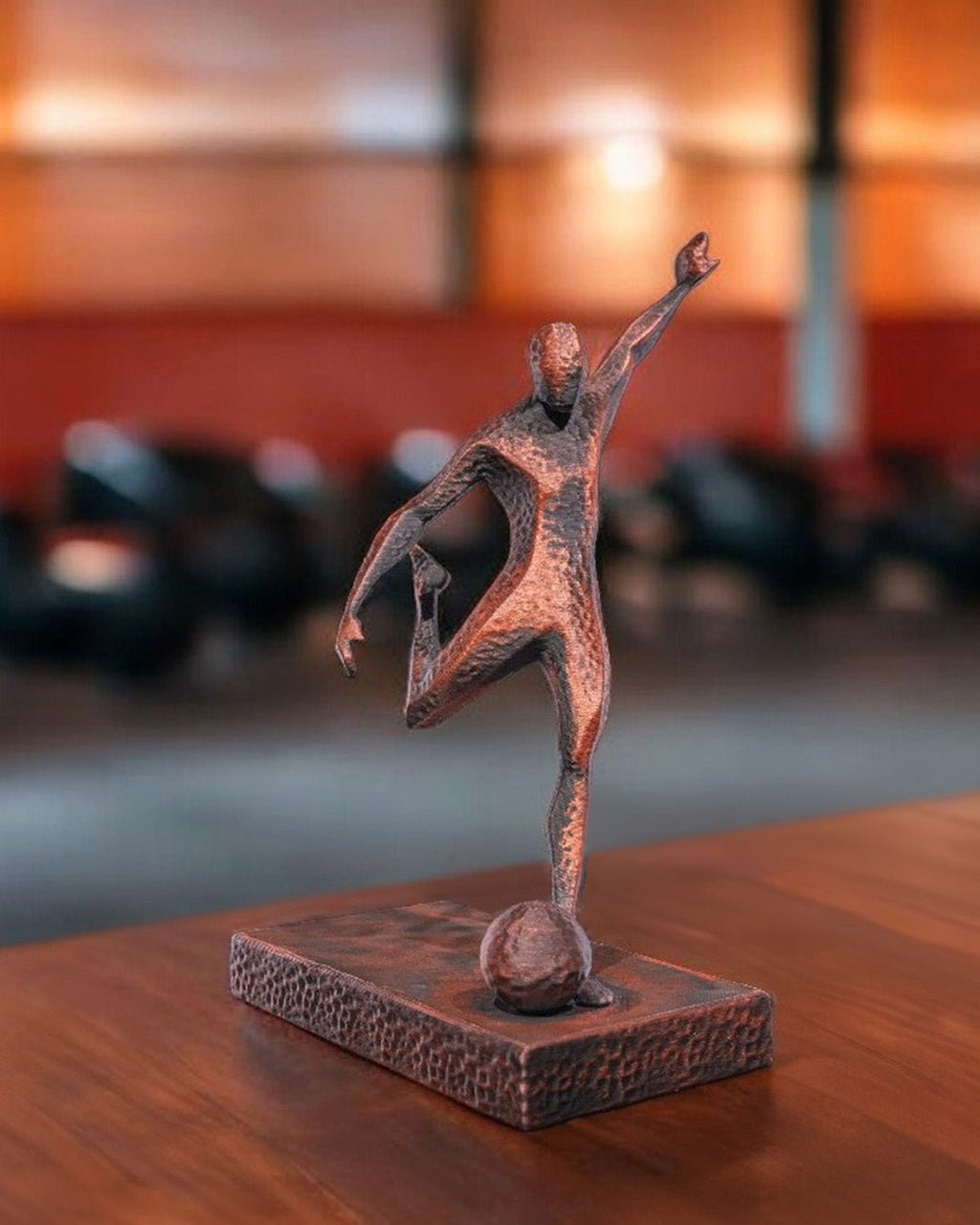 Fußballspieler-Statue in Kupfer-Optik, Frontalansicht auf Holztisch, unscharfer Hintergrund mit Stühlen und roter Wand – Münch3D