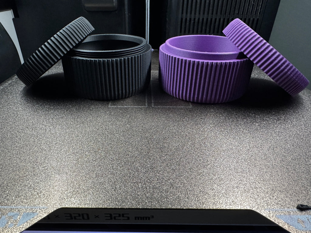 Schwarze und lila geriffelte Aufbewahrungsdose direkt vom 3D-Drucker, beide geöffnet auf dem Druckbett, Druckerrahmen und Display im Hintergrund sichtbar