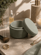 Geöffnete salbeigrüne geriffelte Aufbewahrungsdose auf Marmor-Waschtisch, umgeben von Make-up-Pinsel, goldenen Ringen, Kette, Duftöl-Fläschchen und Seifenspender