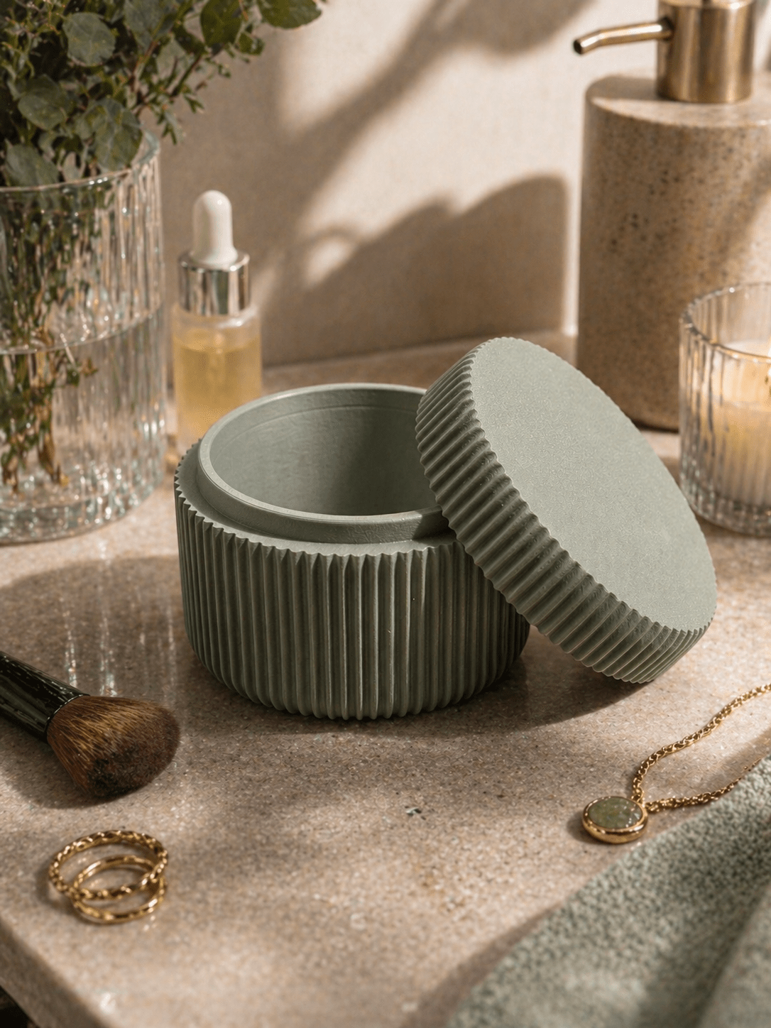 Geöffnete salbeigrüne geriffelte Aufbewahrungsdose auf Marmor-Waschtisch, umgeben von Make-up-Pinsel, goldenen Ringen, Kette, Duftöl-Fläschchen und Seifenspender