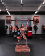 Handbemalte Gewichtheber-Statue in Kupfer-Optik, 3D-Druck im Fitnessstudio auf dunklem Tisch, unscharfer Gym-Hintergrund – Münch3D