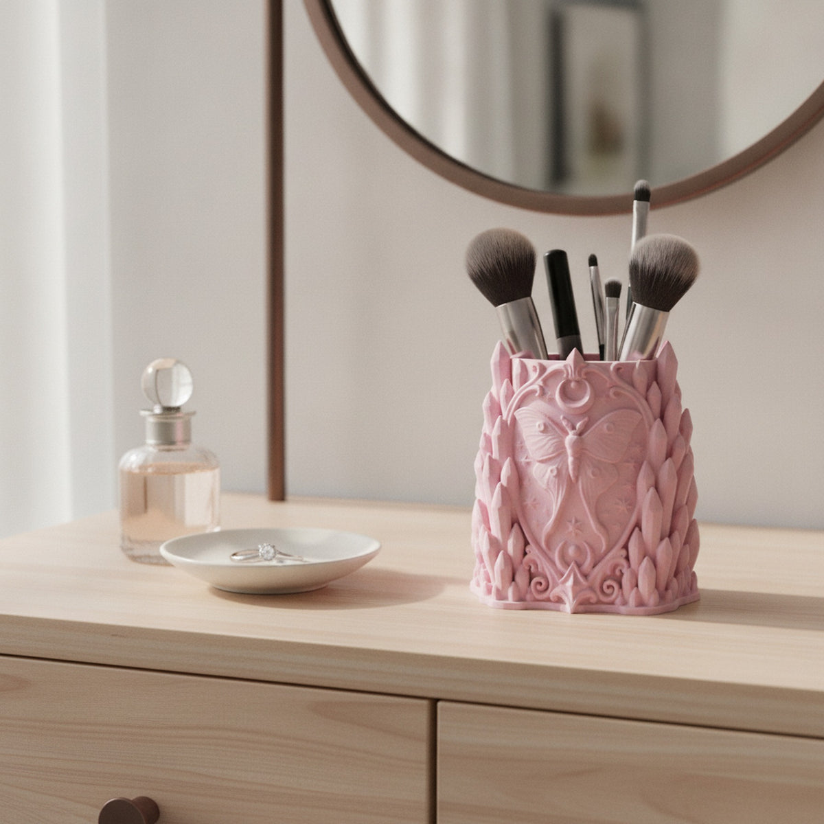 Rosa Kristall-Pinselhalter mit Mottenmotiv auf einer Holzkommode neben Parfümflakon und Ringschale vor einem Spiegel