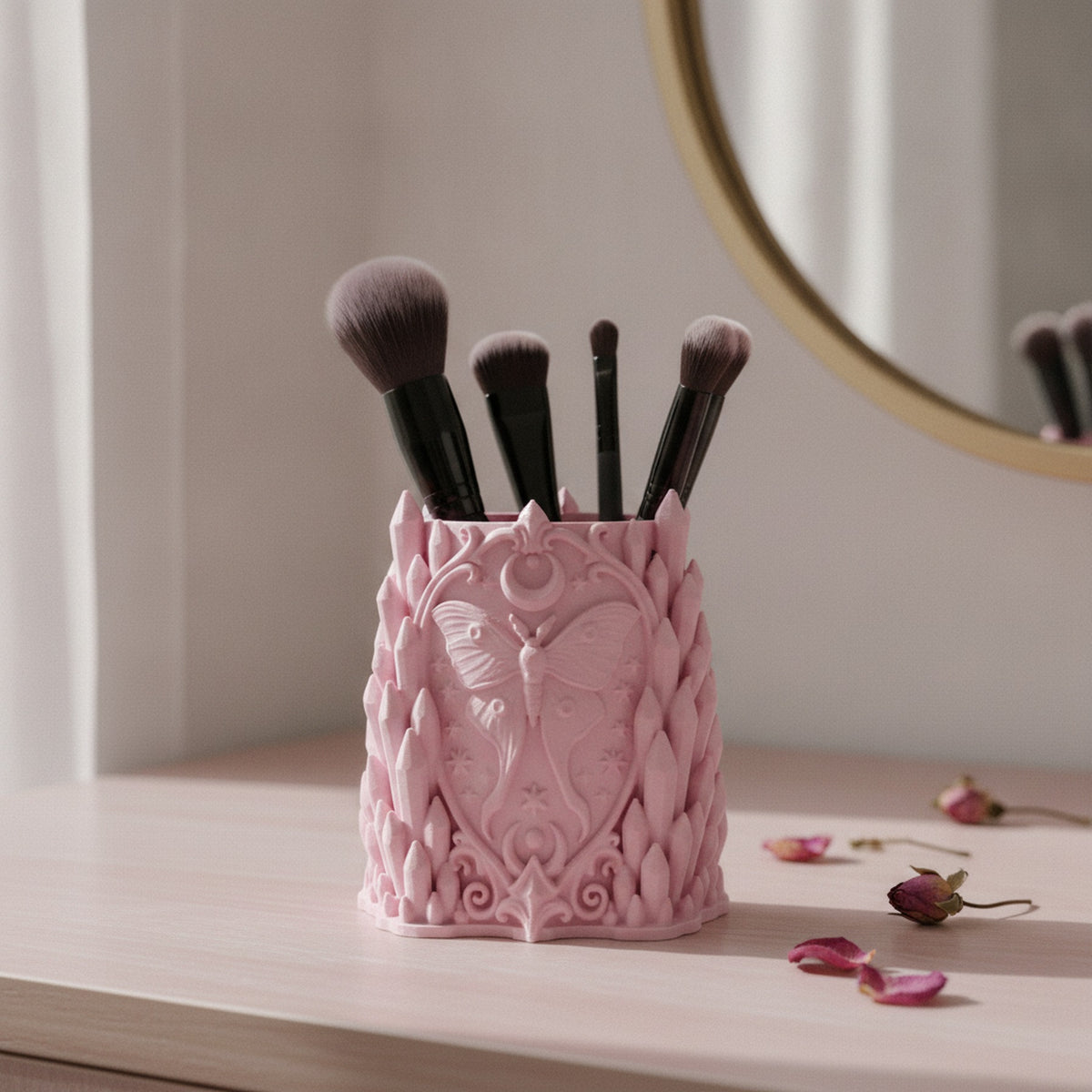 Rosa Gothic Kristall-Pinselhalter mit Luna-Motten-Relief, gefüllt mit Make-up-Pinseln auf einem hellen Schminktisch vor einem runden Spiegel