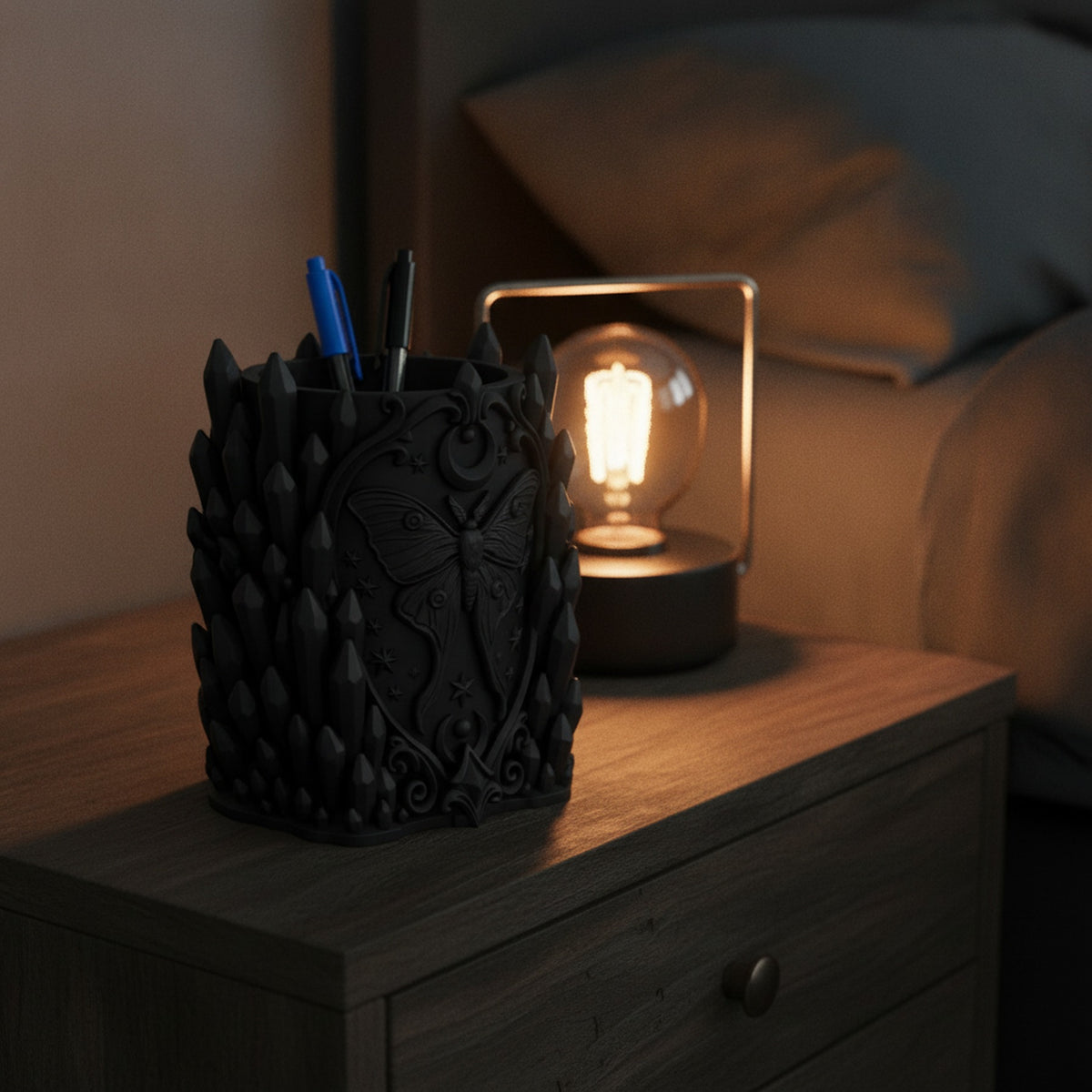 Schwarzer Kristall-Stiftehalter mit Motten-Relief auf einem Nachttisch neben einer warm leuchtenden Nachttischlampe im abendlichen Schlafzimmer