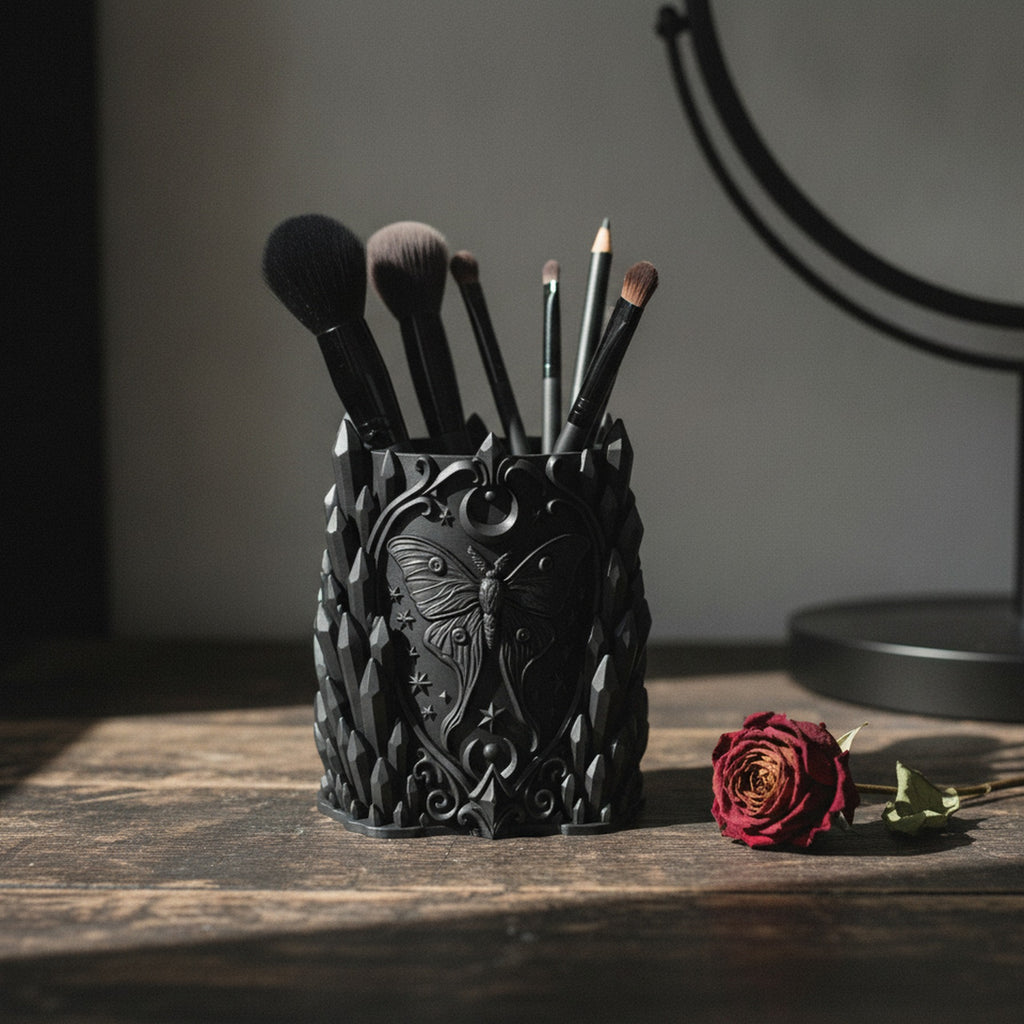 Schwarzer Gothic Kristall-Pinselhalter mit Luna-Motten-Relief, gefüllt mit Make-up-Pinseln auf dunklem Holztisch neben einer getrockneten Rose