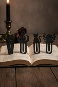 Gothic-Lesezeichen-4er-Set mit Sarg, Mondkrone, Katze und Kristallkugel, alle vier mattschwarz an der Oberkante eines aufgeschlagenen Buches auf einem dunklen Holztisch geklemmt, im Hintergrund eine schwarze Kerze