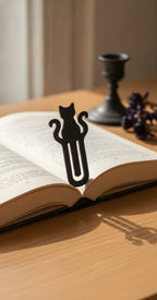 Mattschwarzes Katzen-Lesezeichen mit vollständiger Katzen-Silhouette und geschwungenem Schweif auf einem aufgeschlagenen Buch, Holztisch mit Kerzenständer und getrockneten Blüten im Hintergrund
