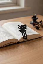 Mattschwarzes Kristallkugel-Lesezeichen mit Funken- und Rauch-Ornament auf der Oberkante eines aufgeschlagenen Buches geklemmt, Holztisch mit Kerzenständer und getrockneten Blüten im Hintergrund