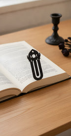Mattschwarzes Dreifacher-Mond-Lesezeichen mit Mondkrone auf der Oberkante eines aufgeschlagenen Buches geklemmt, Holztisch mit Kerzenständer und getrockneten Blüten im Hintergrund