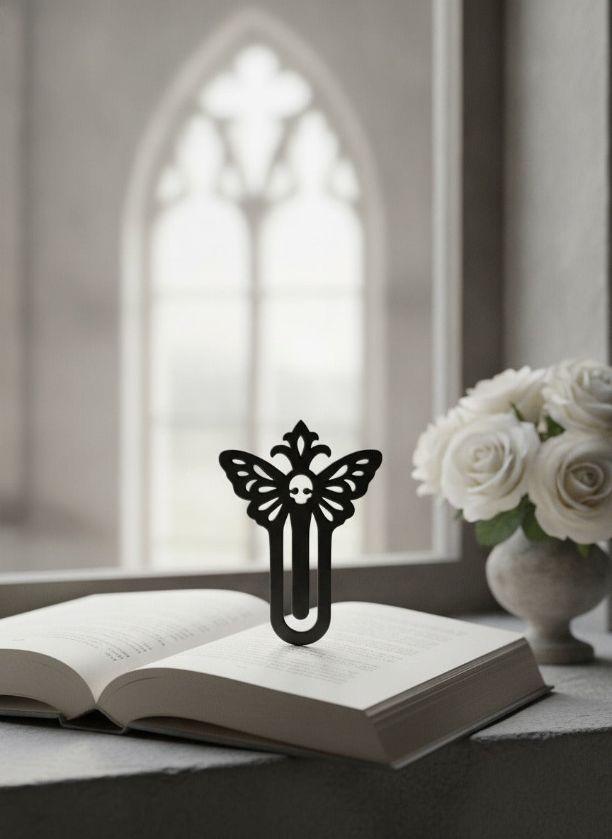 Schwarzes Totenkopf-Falter-Lesezeichen an der Oberkante eines aufgeschlagenen Buches auf einer Fensterbank vor gotischem Kirchenfenster geklemmt, weiße Rosen im Hintergrund