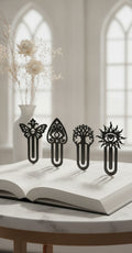 Gothic-Lesezeichen-4er-Set mit Sonnenauge, Mondbaum, Totenkopf-Falter und Amulett-Auge, alle vier mattschwarz an der Oberkante eines aufgeschlagenen Buches auf einem hellen Marmortisch geklemmt, im Hintergrund eine weiße Vase mit getrockneten Blüten