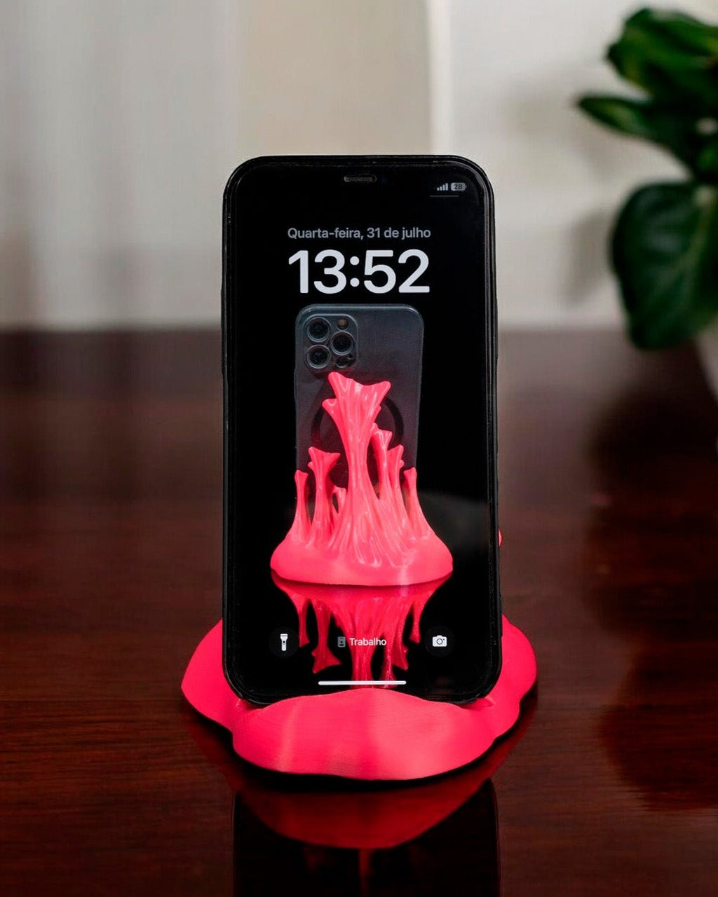 Neonpinker Splash-Handyhalter mit eingestecktem Smartphone auf Holztisch, Frontalansicht mit Pflanze im Hintergrund