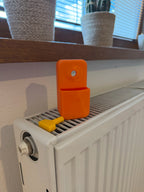 Orange Halterung mit gelbem Entlüftungsschlüssel steht auf einem weißen Heizkörper vor einem Fenster mit Pflanzen