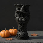 Schwarze Hexen-Kürbis-Vase ohne Blumen mit Jack-o'-Lantern-Gesicht am Sockel, daneben zwei echte Kürbisse und Herbstblätter auf dunkler Fläche