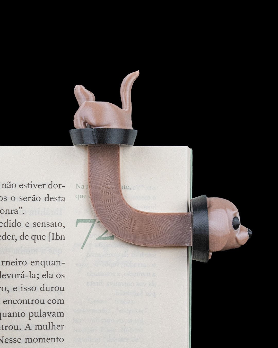 Braunes Hunde-Lesezeichen aus dem 3D-Drucker auf aufgeschlagenem Buch mit lesbarem Text – Hinterteil mit hochgestellten Ohren und Schwanz ragt oben aus den Seiten, Kopf mit Kulleraugen schaut seitlich über die Buchkante