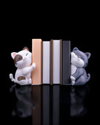 Katzen-Buchstützen-Set: weiß-braune und graue Katzenfigur halten fünf Bücher (pfirsich, weiß, dunkelgrau) zwischen sich, Frontalansicht vor schwarzem Hintergrund