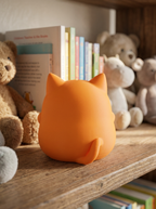 Orangefarbene Katzen-Spardose von hinten auf Holzregal, mehrere Teddybären und Bücherstapel im Hintergrund