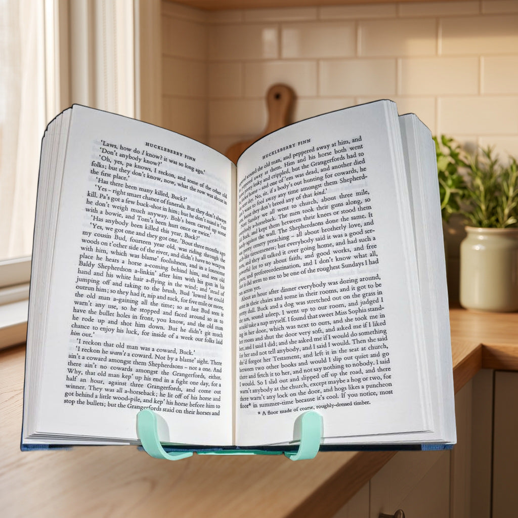 Buchständer mit aufgeschlagenem Buch (Huckleberry Finn) von vorne, Textseiten symmetrisch und gut lesbar. Im Hintergrund Kochbücher und Vorratsdosen auf der Küchenarbeitsplatte.