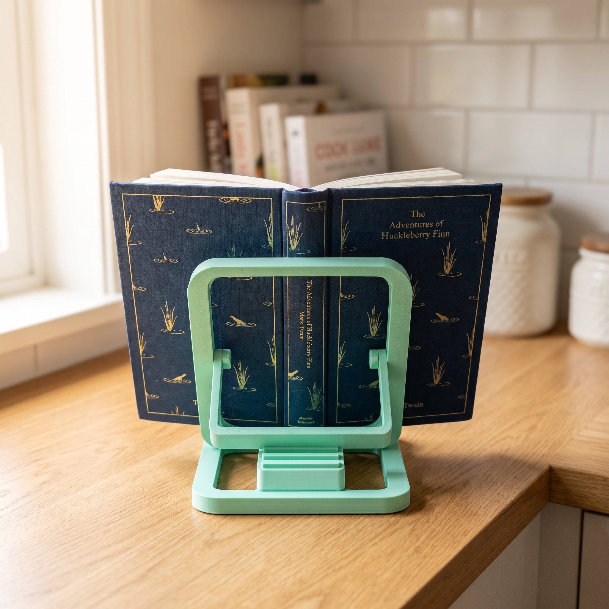 Buchständer mit geschlossenem Buch frontal im Ständer, Covertext „The Adventures of Huckleberry Finn“ und Buchrücken gut lesbar. Symmetrische Ansicht auf der Küchenarbeitsplatte mit Kochbüchern und Vorratsdosen im Hintergrund.