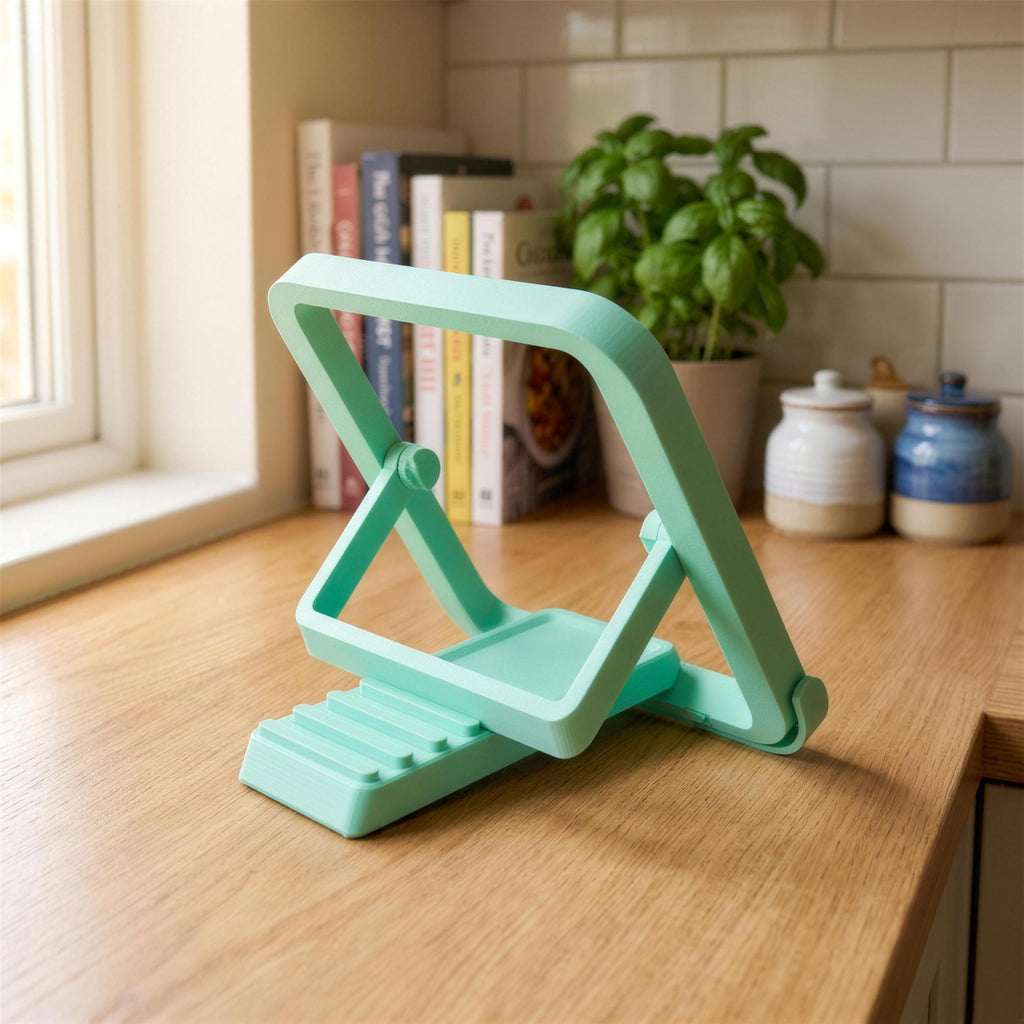 Leerer Buchständer diagonal von der Seite, Klappmechanismus mit Scharnier sichtbar. Im unscharfen Hintergrund Fensterlicht, Kochbücher und eine Topfpflanze auf der Arbeitsplatte.
