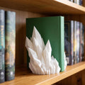 Weiße Kristall-Buchstütze mit spitzen, facettierten Kristallzacken steht auf einem Holzregal vor einem aufrecht stehenden grünen Buch, links und rechts weitere Bücher mit bunten Buchrücken.